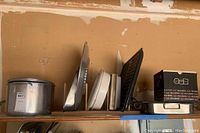 Wide view of shelf showing large metal pot, baking pans, broiling pan, white plastic trays and veggie slicer.