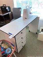 Photo of white sewing table with black edges, fold-up design, and sewing machine on top (machine not included). Shows the full table.