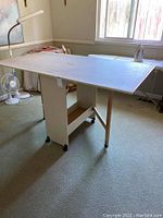 Front side angle view of the beige adjustable utility sewing table holding a large white cutting board with grid measurements, showing collapsible legs and caster wheels.
