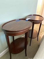 Photo of two vintage wooden side tables with oval tops and lower shelves. Dark wood finish, tapered legs. Used condition.