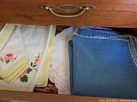 Photo of embroidered napkins, white and yellow with floral designs and blue linens with patterned edges