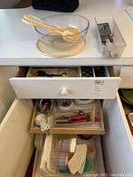 Photo of a glass salad bowl with two yellow serving utensils on top, a container with metal and plastic kitchen gadgets to the right, and open drawers showing white measuring spoons and cups, Tupperware lids and containers inside.