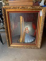 Front view of vintage painting featuring woman in blue dress playing harp, ornate gilded frame visible