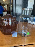 Brown glazed ceramic pot with lid and clear glass beverage dispenser on wooden surface, kitchen background