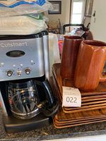 Cuisinart stainless steel coffee maker with glass carafe, glass pot handle, and power cord wrapped around it, sitting next to wooden square plates and brown wooden mugs. Items on a kitchen counter.