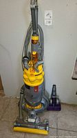 Full front view of the Dyson upright vacuum cleaner with yellow and gray body, hose wrapped around, and power cord.