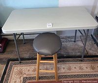 Full view of rectangular plastic folding table with wooden stool placed underneath on a patterned rug.