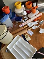 Multiple Tupperware containers, rolling pins, measuring spoons, cutting boards, hand press, glass measuring cups, and scooping spoons arranged on a wooden surface.