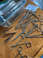 Photo showing stainless steel medical instruments including multiple hemostats, tweezers, scissors, and a glass sterilizing vessel with metal insert lid on a wooden surface.