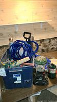 Photo showing a blue bin with coiled blue Christmas string lights, black lantern with green top, propane fuel canisters, bird feeders, and teapot items on a wood table.
