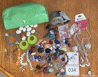 Green zipper pouch, assorted polished stones, shell jewelry pieces, and small crafting components laid out on wood surface.