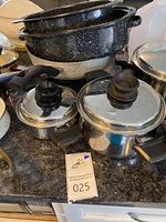 Photo showing black speckled roasting pans stacked with various stainless steel pots and pans with lids on a countertop.