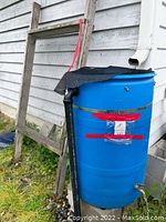 Blue plastic rain barrel outside next to a house, approximately 37 inches tall and 22 inches diameter, covered by a black mesh sieve/barrel cover secured on top. The barrel rests on cinder blocks and is near a rain gutter downspout.