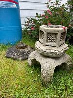 Concrete garden pagoda with multiple tiers and geometric cutouts placed on grass next to flowers and the side of a building, showing weathering and moss.