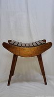 Front view of the wooden stool showing curved slatted seat and four angled legs with natural wood finish and visible wear.
