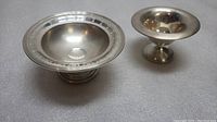 Two sterling silver serving dishes on a grey surface, one larger with decorative rim, one smaller with plain rim.