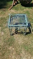 Top-down view of the square outdoor wrought iron table with glass top on grass showing frame and glass surface.
