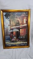 Full view of framed oil painting showing street cafe scene with tables, chairs and umbrellas on street sidewalk by building.