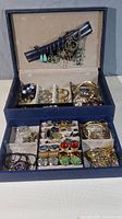 Top view of open blue jewelry box showing earrings hung on lid and various jewelry compartments filled with rings, brooches, bracelets, and cufflinks.