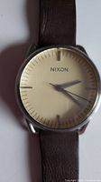 Close-up image of the Nixon men's watch showing beige round dial with silver hour markers and hands, and a brown leather strap.