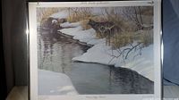 Framed print showing three wolves on a snowy riverbank with a river reflecting the winter landscape.