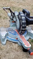 Full side view of the sliding compound miter saw on grass, showing the handle, blade guard, base with angle markings, and sliding rails.