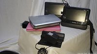 Three portable DVD players stacked along with black power adapters and cords in front. Two players with black screens behind, one silver player on top, and pink case visible underneath.