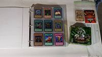 Binder open showing 9 Yu-Gi-Oh! cards in clear sleeves with a green Yu-Gi-Oh! box and some loose cards sealed in a plastic bag, plus a rule book
