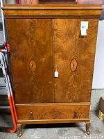 Front closed view of the antique solid wood armoire showing wood grain and inlay details on doors and drawer, with hanging tags.