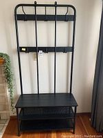 Full view of the black metal coat rack with shoe storage bench, showing the four central vertical bars, top horizontal rows with hooks, and bench with two shelves.