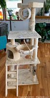 Front right angle view of the beige tri-level cat tree condo revealing platforms, side hammock, circular tunnel, ladder, and enclosed cubby.