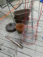 Photo showing two brown plastic deck railing planters stacked, two terra cotta pots, and a heavy brown umbrella stand base on a wooden decking surface, also visible metal tomato cages and packing sticks.
