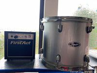 Compact black and blue First Act amplifier with control knobs on top, placed next to the drum.