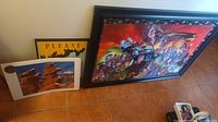 Grouped photo showing one large framed Justice League poster with colorful comic art, one smaller framed yellow and brown "Please Keep The Park Clean" poster, and three white-bordered unopened photos of Garden of the Gods leaning against a wall on tile floor.