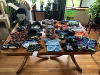 Wide image showing Frankenstein Build-A-Bear plush seated with numerous clothes and accessories laid out on wooden table including pajamas, shirts, hats and jerseys for dolls.