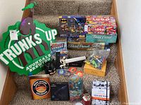 Photo showing the Avondale Brewery tin sign, several board games including Trivial Pursuit, Logo, and Batman puzzle, a bag with toy cars, and small collectible items arranged on carpeted stairs.