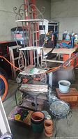 Overall view of iron folding plant stand, various pots, gardening supplies, and small tables in garage setting