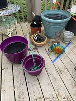 Photo of assorted outdoor planters including large blue ceramic-style planter, purple plastic pots, gray pots, black nursery pots, and hanging moss basket with hummingbird feeder and glass birdbath on deck.