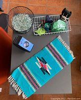 Top view of lot showing southwestern woven rug, various decorative glass rocks, batman plush, yo-yo, starry night projector, wire racks.