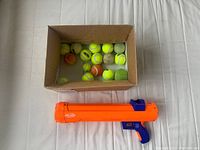 Nerf Dog tennis ball launcher placed in front of a cardboard box holding multiple tennis balls of sizes suitable for dogs.
