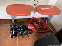 Nike orange padded exercise bench with silver metal legs, hand weights, adjustable dumbbell bars, resistance band, and deflated red stability ball arranged on tile floor.