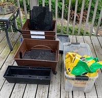 Three brown City Pickers raised garden grow boxes with caster wheels outdoors on wooden deck, showing perforated grid platforms and vertical watering pipes inside. A clear Rubbermaid tote without a lid contains one unopened and one partial bag of Miracle Gro potting mix. Several black plastic rectangular planters are also present.