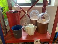 Photo showing the lot items including the red metal watering can, glass and plastic terrarium planters with rope hangers, milk glass horse head vase, gold-toned planter pots, ceramic vases, self-watering globes, and coffee mug all arranged on a red shelf