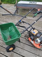 Powermate gas tiller connected to Scotts green seed spreader on wooden deck, showing overall lot condition and setup.
