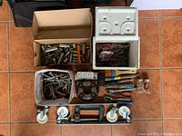 Top-down view of assorted tools and hardware in boxes and containers including sockets, drill bits, wrenches, and paintbrushes on tiled floor.