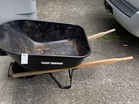 Side view of True Temper metal wheelbarrow showing black metal tub with some rust, wooden handles, and single wheel.