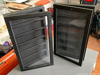Two black wooden and glass coin display cases with glass fronts, standing open and empty on a surface showing interior with black velour lining and grooved shelves
