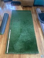 Top view photo of larger roll of plush artificial grass on wooden floor with measuring stick showing length and width approximately 91" by 46".