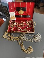 Photo showing multiple pieces of vintage costume jewelry including chain belts, bracelets, earrings, necklaces, and pendants, displayed open on a gray surface near the red velvet-lined jewelry case.