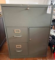 Front view of vintage grey metal cabinet showing closed compartments, two filing drawers on left and door on right, lock on upper flip-down desk compartment.